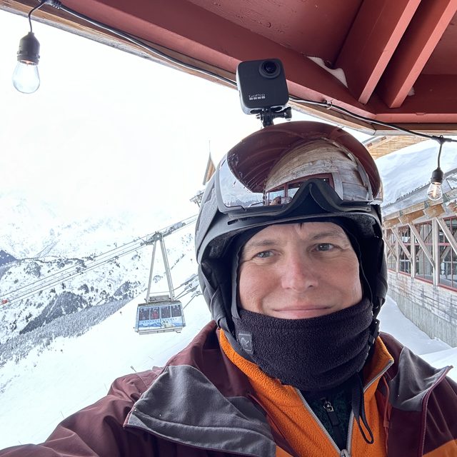 Seth Baker in a ski helmet at the top of a mountain, an aerial tram cabin and snowy ridgeline behind him